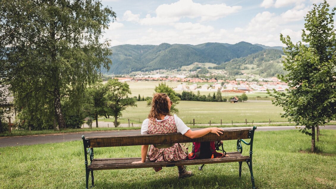 Eine Person sitzt auf einer Bank und blickt auf eine malerische Landschaft mit Bergen und einem Dorf im Hintergrund. Der Himmel ist bewölkt und die Umgebung ist grün und einladend. | © TV Hochsteiermark, nicoleseiser.at