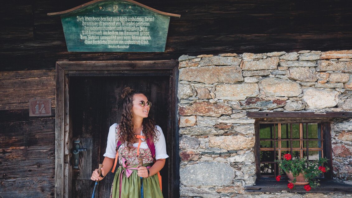 Eine Frau steht vor einem traditionellen Steinhaus und trägt eine Tracht. Im Fenster blühen rote Pflanzen, und über der Tür hängt ein Schild mit Text. | © TV Hochsteiermark, nicoleseiser.at