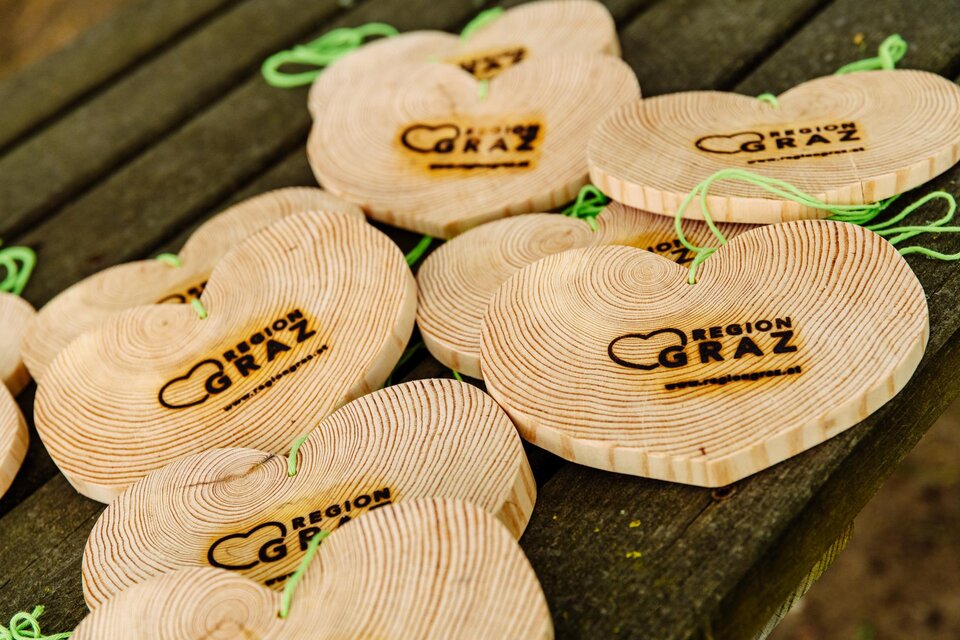 Holzherzen mit dem Logo "Region Graz" liegen auf einem Tisch. Sie sind aus naturbelassenem Holz und haben grüne Bänder zum Aufhängen. | © Mias Photoart