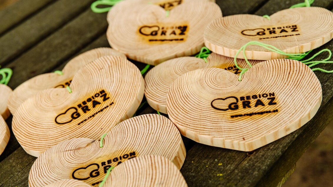 Holzherzen mit dem Logo "Region Graz" liegen auf einem Tisch. Sie sind aus naturbelassenem Holz und haben grüne Bänder zum Aufhängen. | © Mias Photoart