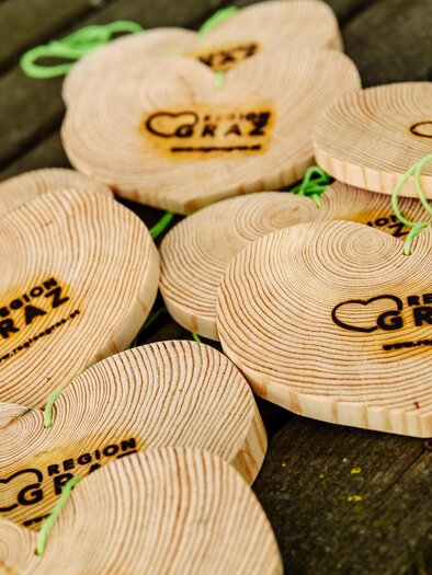 Wooden hearts with the "Region Graz" logo are laid out on a table. They are made from untreated wood and have green ribbons for hanging. | © Mias Photoart