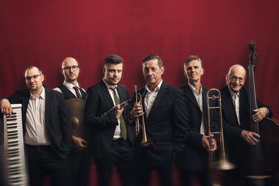 Eine Gruppe von sechs Musikern steht vor einem roten Hintergrund. Sie halten verschiedene Instrumente wie Trompete und Posaune und tragen formelle Kleidung. | © Dixie Hot Seven