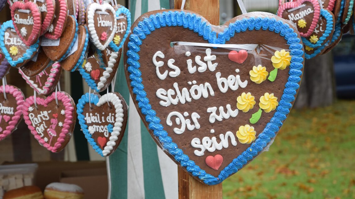 Ein herzförmiges Lebkuchen-Schild mit der Aufschrift "Es ist schön bei Dir zu sein" und bunten Verzierungen. Im Hintergrund hängen weitere süße Herzschilder. | © Marktgmeinde Niklasdorf
