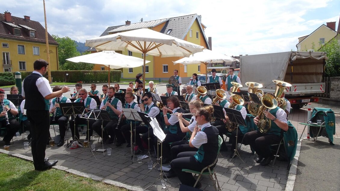 Eine Blaskapelle spielt im Freien unter Sonnenschirmen. Im Hintergrund sind bunte Häuser und ein Lastwagen zu sehen. | © Marktgemeinde Niklasdorf