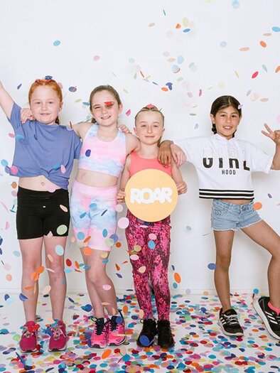 Vier fröhliche Kinder stehen vor einem weißen Hintergrund und feiern mit bunten Konfetti. Sie tragen sportliche Kleidung und lächeln in die Kamera.