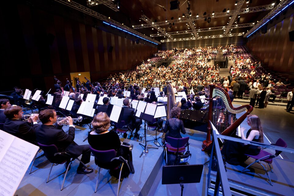Ein Orchester spielt vor einem großen Publikum in einem modernen Konzerthaus. Die Musiker sitzen auf der Bühne und konzentrieren sich auf ihre Instrumente. | © Graz Tourismus-Harry Schiffer