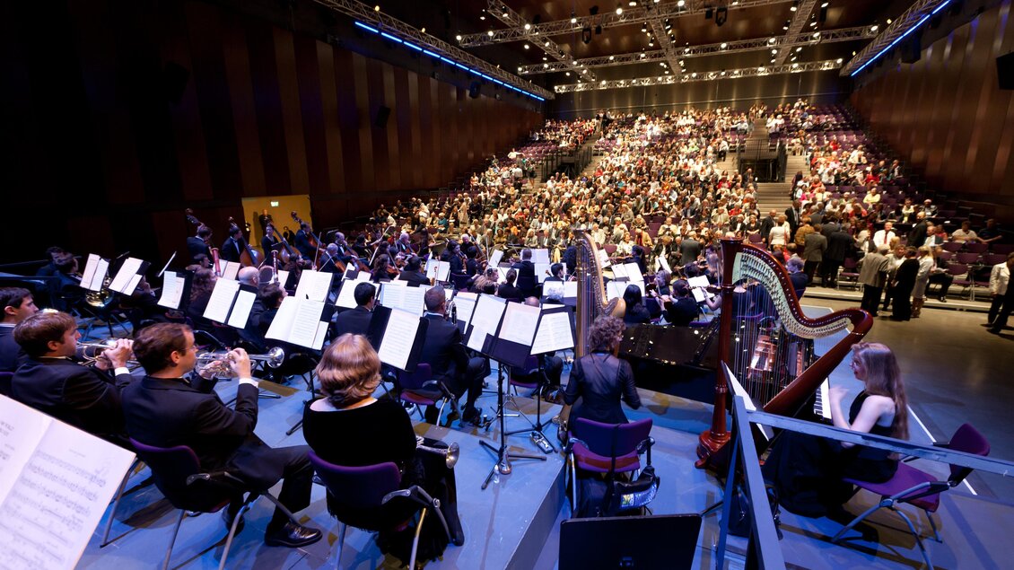Ein Orchester spielt vor einem großen Publikum in einem modernen Konzerthaus. Die Musiker sitzen auf der Bühne und konzentrieren sich auf ihre Instrumente. | © Graz Tourismus-Harry Schiffer