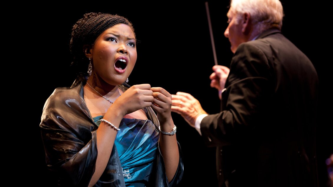 Eine Sängerin in einem hübschen blauen Kleid singt leidenschaftlich auf der Bühne. Im Hintergrund steht ein Dirigent, der die Aufführung leitet. | © Graz Tourismus-Harry Schiffer