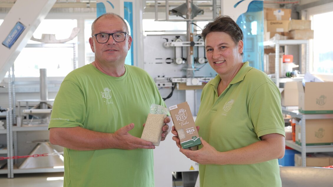 Zwei Personen in grünen Shirts stehen in einer Produktionsumgebung. Sie halten Verpackungen in den Händen und lächeln freundlich. | © SteirerREISbyFuchs