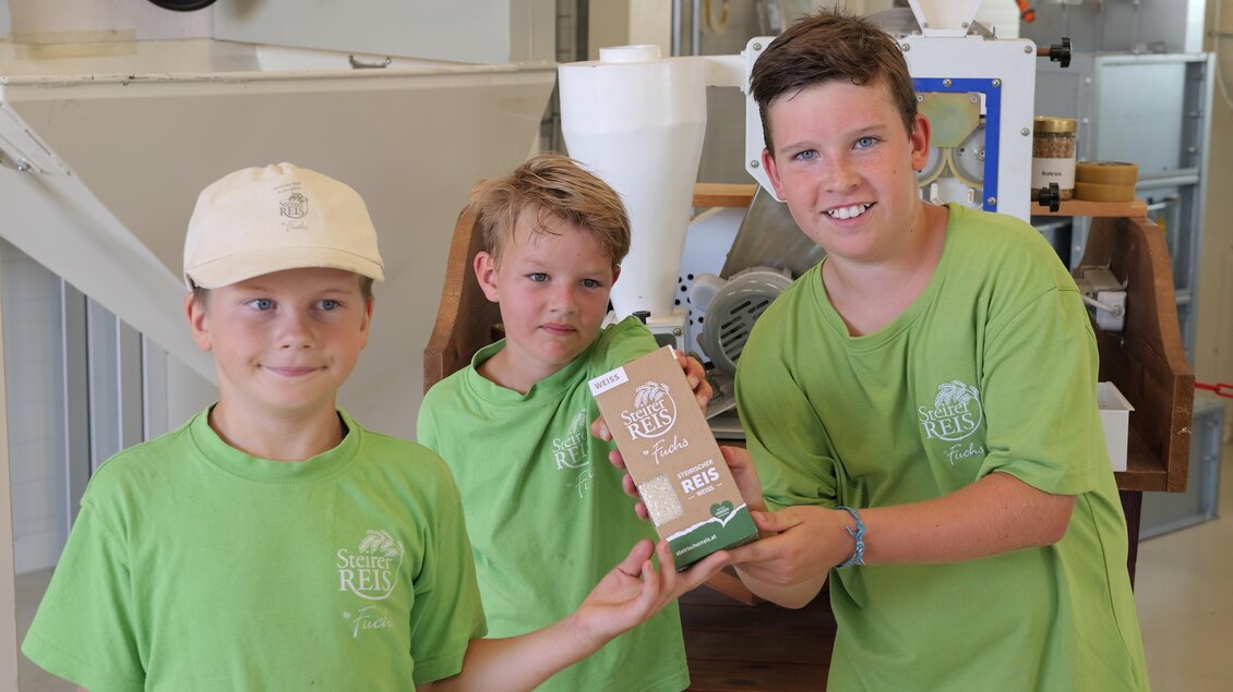 Drei Kinder in grünen T-Shirts halten eine Verpackung in der Hand. Im Hintergrund sind Maschinen und Werkzeuge zu sehen. | © SteirerREISbyFuchs