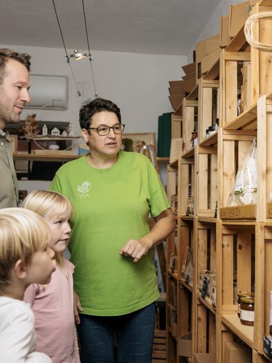 Eine Familie schaut interessiert auf Produkte in einem Laden. Die Verkäuferin erklärt den Kunden die Artikel in den Holzkisten. | © Vulkanland Knittelfelder Christian