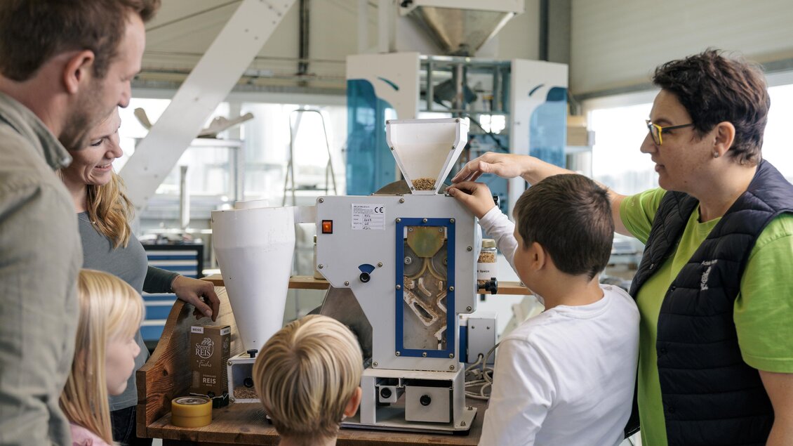 Eine Gruppe von Erwachsenen und Kindern schaut auf eine Maschinenanlage zur Verarbeitung von Getreide. Eine Person erklärt den Aufbau und die Funktionsweise der Maschine. | © Vulkanland Knittelfelder Christian