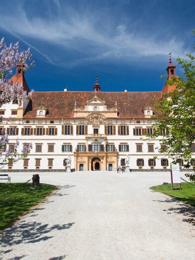 Ein beeindruckendes Schloss mit einer prächtigen Fassade und roten Dächern. Umgeben von grünem Rasen und blühenden Bäumen unter einem klaren blauen Himmel. | © Graz Tourismus - Harry Schiffer