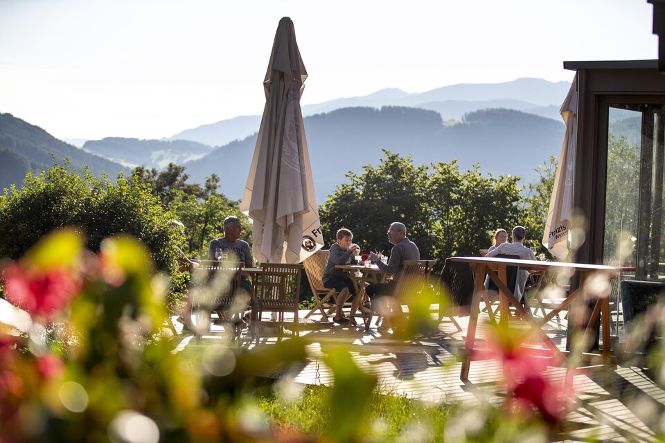 Ein schöner Außenbereich mit Tischen und Sesseln, wo Menschen in der Sonne sitzen. Im Hintergrund sind Berge und ein blauer Himmel zu sehen. | © TV Murau