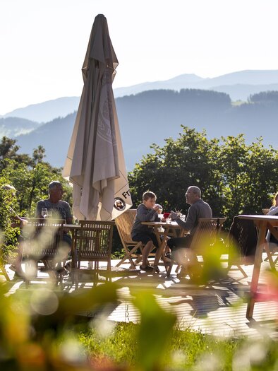 A beautiful outdoor area with tables and chairs, where people sit in the sun. In the background, mountains and a blue sky can be seen. | © TV Murau