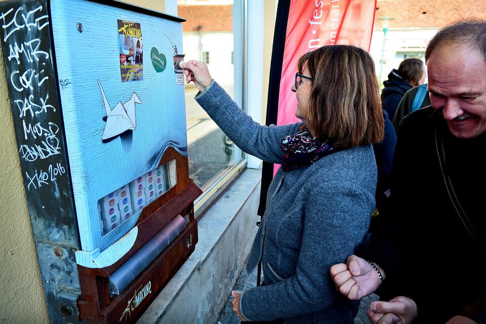 Eine Frau interagiert mit einem künstlerischen Automaten an einer Wand. Hinter ihr steht ein Mann, der lächelt und die Szene beobachtet. | © Strassegger