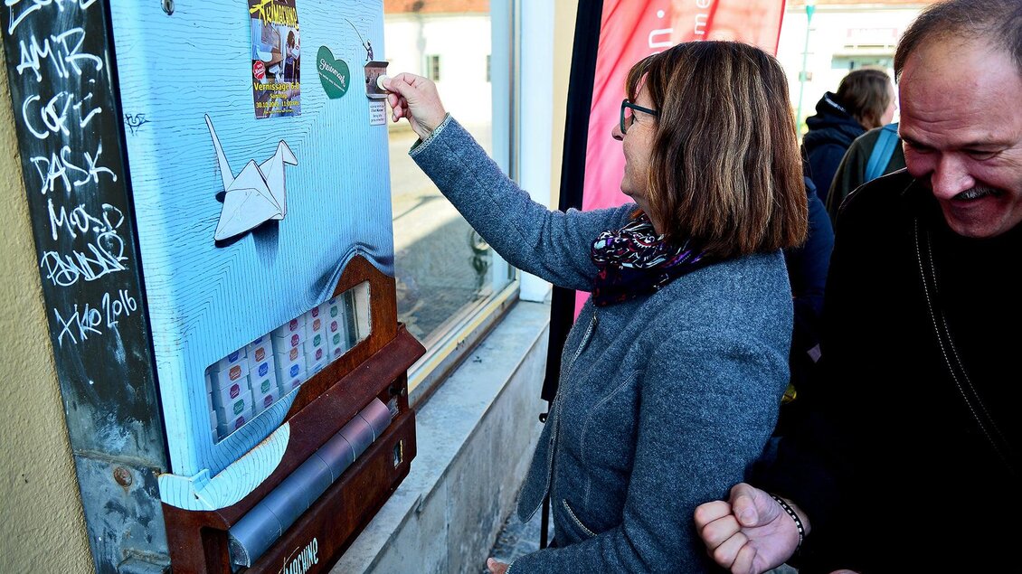 Eine Frau interagiert mit einem künstlerischen Automaten an einer Wand. Hinter ihr steht ein Mann, der lächelt und die Szene beobachtet. | © Strassegger