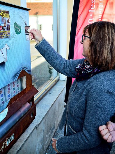 Eine Frau interagiert mit einem künstlerischen Automaten an einer Wand. Hinter ihr steht ein Mann, der lächelt und die Szene beobachtet. | © Strassegger