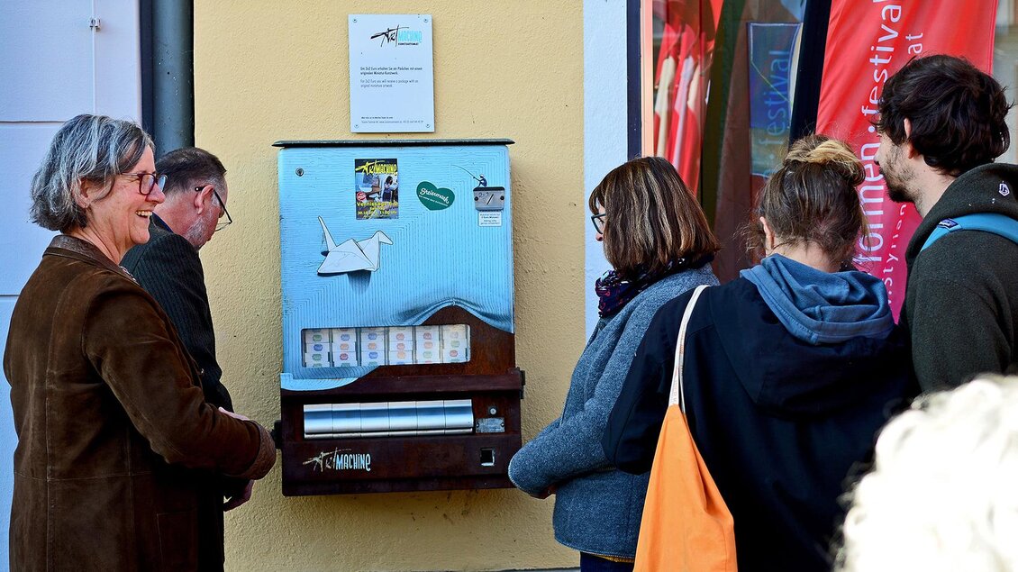 Eine Gruppe von Menschen steht vor einem Verkaufsautomaten an einer Wand. Der Automat bietet verschiedene Produkte zum Verkauf an. | © C. Strassegger