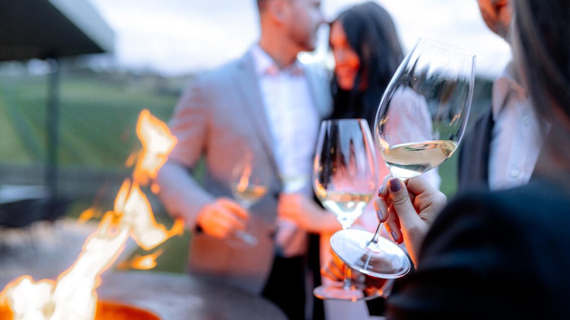 Eine elegante Feier im Freien mit fröhlichen Menschen, die Wein in Gläsern halten. Im Hintergrund ist ein Lagerfeuer zu sehen, das eine gemütliche Atmosphäre schafft. | © Ratscher Landhaus