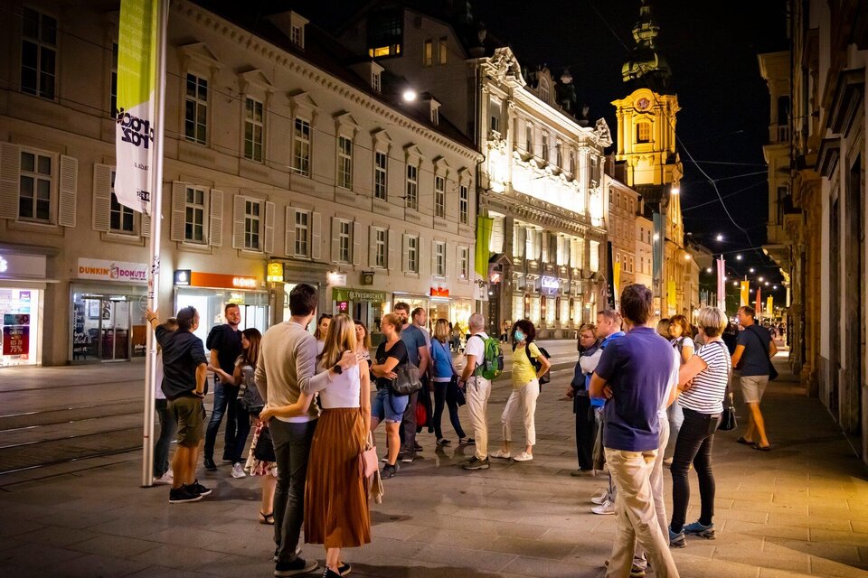 Eine Gruppe von Menschen steht nachts auf einer belebten Straße, umgeben von historischen Gebäuden. Die Straßenbeleuchtung schafft eine einladende Atmosphäre. | © Graz Tourismus - Werner Krug