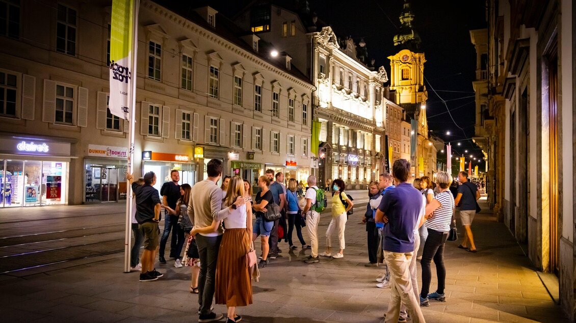 Eine Gruppe von Menschen steht nachts auf einer belebten Straße, umgeben von historischen Gebäuden. Die Straßenbeleuchtung schafft eine einladende Atmosphäre. | © Graz Tourismus - Werner Krug