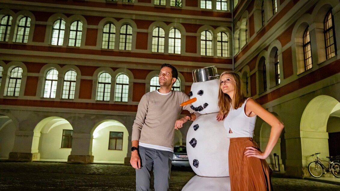 Ein Mann und eine Frau posieren lässig neben einem Schneemann in einem Innenhof. Die Architektur im Hintergrund ist historisch und beleuchtet. | © Graz Tourismus - Werner Krug