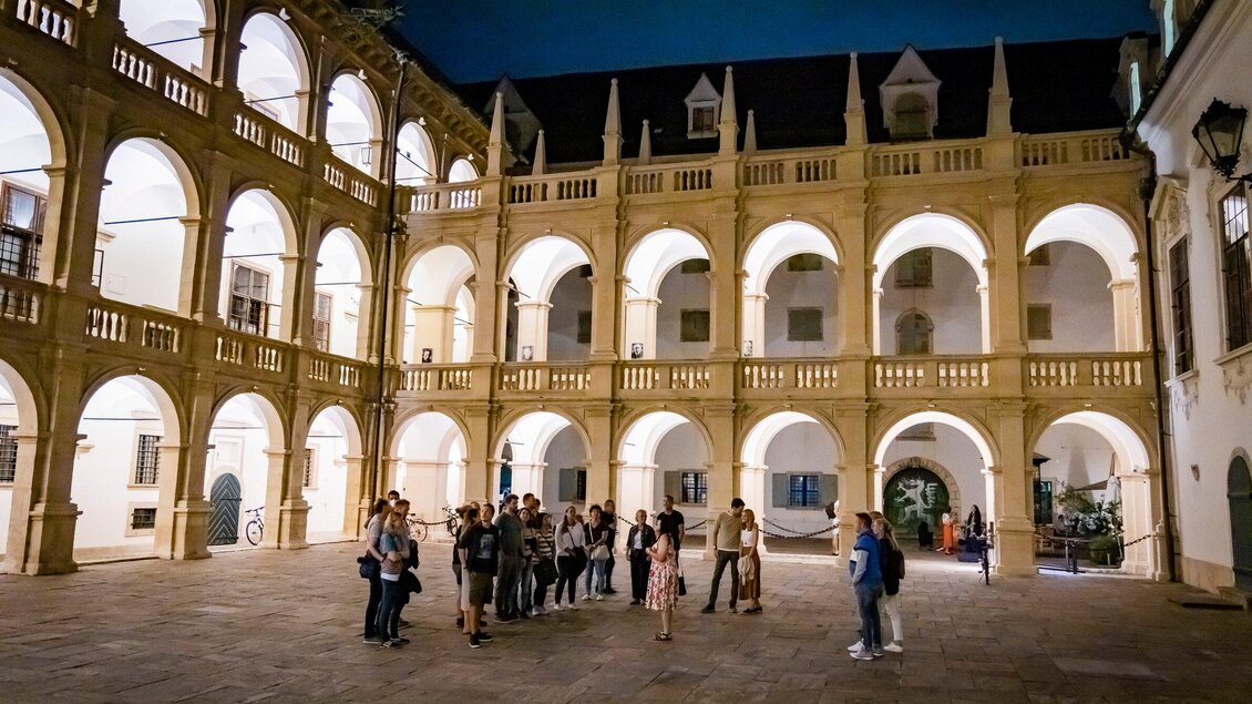 Eine Gruppe von Menschen steht in einem historischen Innenhof mit hohen, bogenförmigen Säulen. Der Abendhimmel leuchtet über dem alten Gebäude. | © Graz Tourismus - Werner Krug