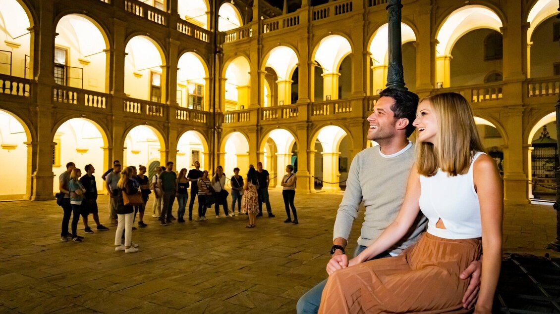 Ein romantisches Paar sitzt in einem erleuchteten Innenhof. Im Hintergrund stehen andere Besucher und genießen die Atmosphäre. | © Graz Tourismus - Werner Krug