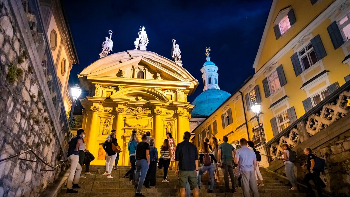 Eine auffällige Kirche mit statuenverzierten Fassaden leuchtet golden in der Nacht. Menschen versammeln sich auf den Treppen und genießen die Abendstimmung. | © Graz Tourismus - Werner Krug