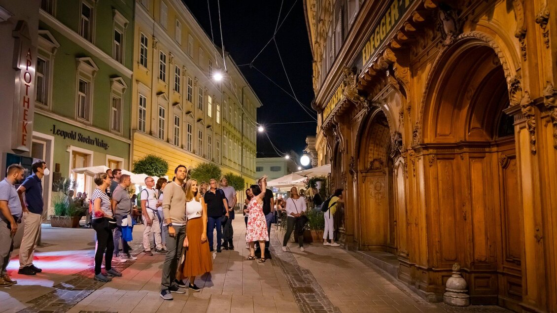Eine belebte Straßenansicht bei Nacht mit mehreren Personen. Im Vordergrund ist ein kunstvoll geschnitztes Holzelement zu sehen. | © Graz Tourismus - Werner Krug