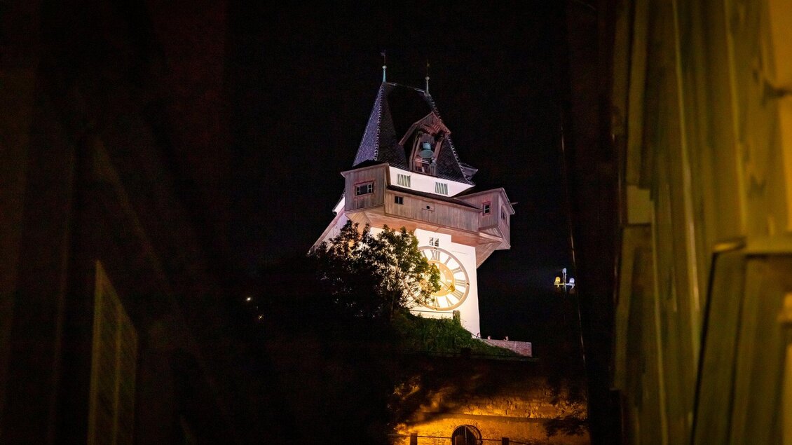 Ein historischer Uhrturm beleuchtet in der Nacht. Das Gebäude ist von dunklen Gassen umgeben und strahlt eine charmante Atmosphäre aus. | © Graz Tourismus - Werner Krug