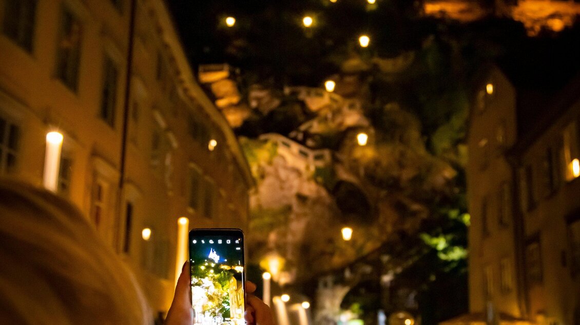 Eine Person hält ein Smartphone und fotografiert eine beleuchtete Felslandschaft bei Nacht. Umgeben von historischen Gebäuden und stimmungsvoller Beleuchtung ist die Szenerie festlich und einladend. | © Graz Tourismus - Werner Krug