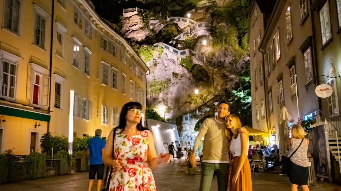Eine malerische Nachtansicht einer Stadtstraße mit bunten Gebäuden. Eine Gruppe von Menschen steht zusammen und genießt die Atmosphäre. | © Graz Tourismus - Werner Krug