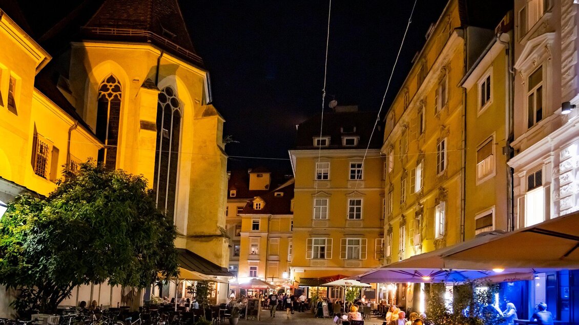 Eine bezaubernde Nachtansicht eines Platzes mit beleuchteten Gebäuden und einem belebten Restaurant. Die Atmosphäre ist einladend und lebhaft. | © Graz Tourismus - Werner Krug