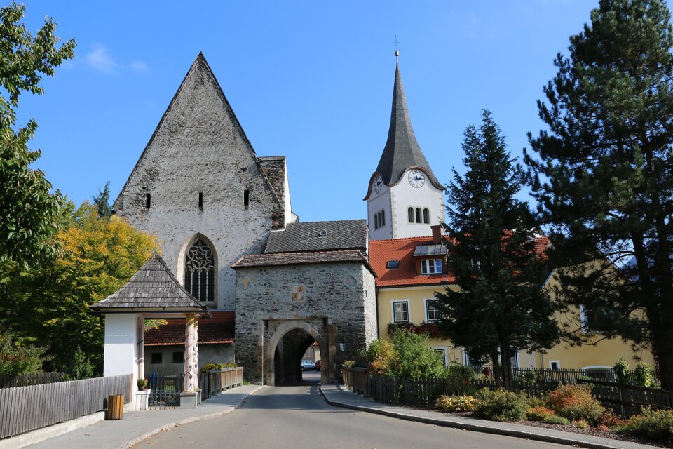 Ein historisches Gebäude mit einem hohen Turm und einem mittleren Bogen. Umgeben von Bäumen und Häusern in einer ruhigen Landschaft.