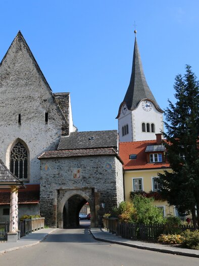 A historic building with a tall tower and a medium arch. Surrounded by trees and houses in a quiet landscape.