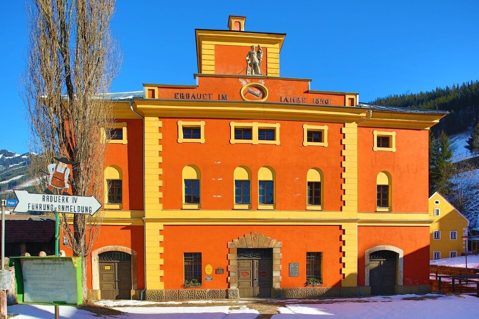 Ein buntes, historisches Gebäude mit einer auffälligen Fassade in Orange und gelb. Im Vordergrund sind Schneeflächen und eine Schilderwand sichtbar. | © Foto Freisinger