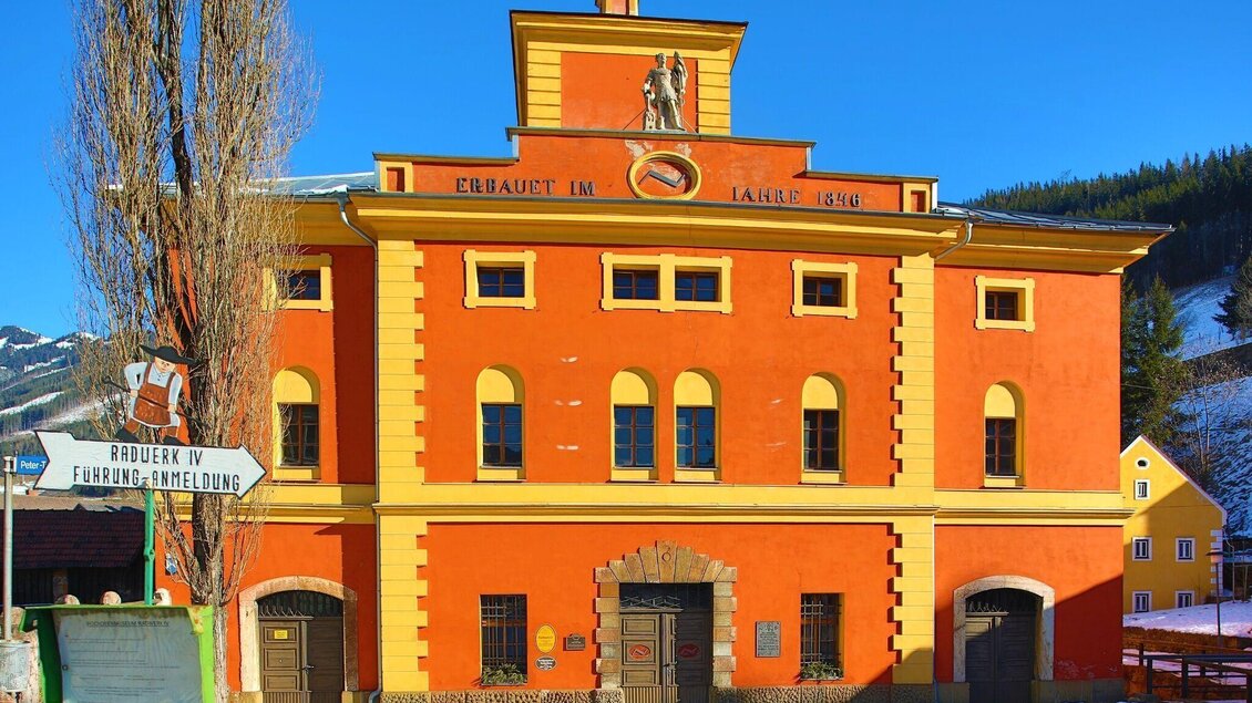 Ein buntes, historisches Gebäude mit einer auffälligen Fassade in Orange und gelb. Im Vordergrund sind Schneeflächen und eine Schilderwand sichtbar. | © Foto Freisinger