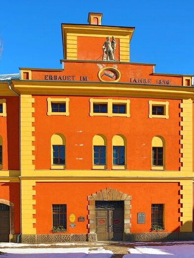 A colorful, historic building with a striking façade in orange and yellow. In the foreground, snow-covered areas and a wall of signs are visible. | © Foto Freisinger