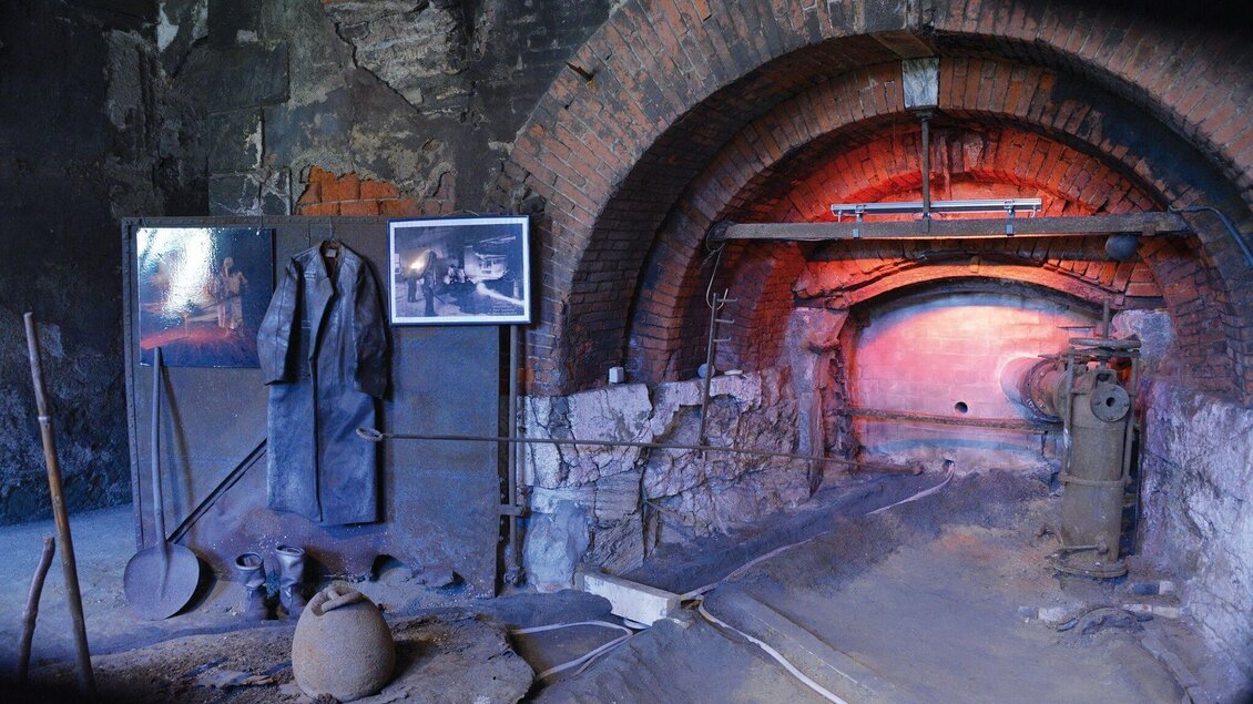 Eine alte, industrielle Innenansicht mit Backsteinbögen und verblassten Fotos an der Wand. Im Vordergrund liegen Werkzeuge und ein Tontopf auf dem Boden. | © RAdwerk IV