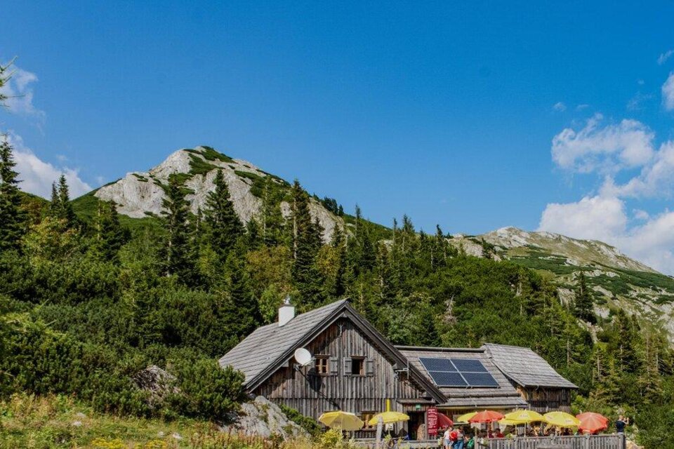 Eine Berghütte umgeben von grünen Bäumen und majestätischen Bergen. Der Himmel ist klar mit einigen Wolken. | © Günther Krenn