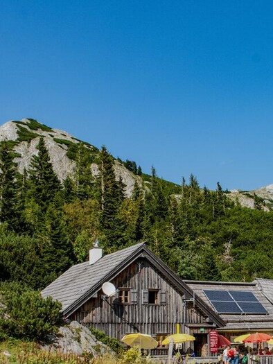 Eine Berghütte umgeben von grünen Bäumen und majestätischen Bergen. Der Himmel ist klar mit einigen Wolken. | © Günther Krenn