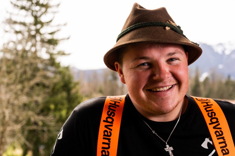 Ein junger Mann mit einem Trachtenhut lächelt in die Kamera. Er trägt ein schwarzes T-Shirt und eine orangefarbene Latzhose.