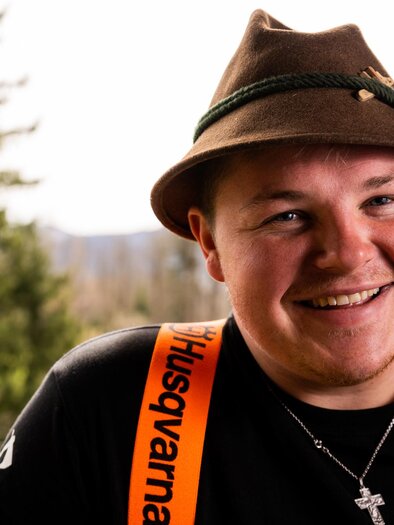Ein junger Mann mit einem Trachtenhut lächelt in die Kamera. Er trägt ein schwarzes T-Shirt und eine orangefarbene Latzhose.