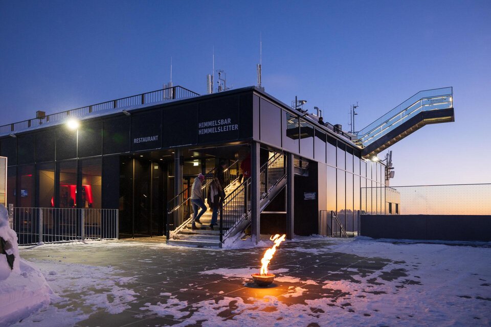 Ein modernes Restaurant auf einem Berg mit Schneedecke. Im Vordergrund brennt eine Flamme, während die Sonne untergeht. | © Martin Huber Adventamdachstein