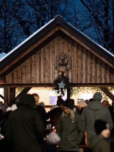 Ein festlicher Weihnachtsmarkt mit beleuchteten Holzständen und vielen Menschen. Die Umgebung ist von winterlicher Atmosphäre und Schnee geprägt. | © Achromatic photography