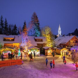 Ein festlicher Weihnachtsmarkt bei Nacht, erleuchtet von bunten Lichtern. Menschen genießen die festliche Stimmung um ein geschmücktes Dorf. | © Martin Huber