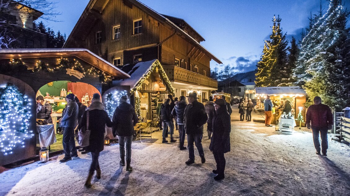 Ein festlicher Weihnachtsmarkt in einer malerischen Winterlandschaft. Menschen schlendern durch den Schnee, umgeben von schimmernden Lichtern und bunten Ständen. | © Gerhard Pilz