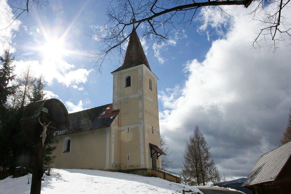 Eine alte Kirche mit einem spitzen Turm steht in einer schneebedeckten Landschaft. Der Himmel ist teils bewölkt, und die Sonne scheint durch die Wolken.